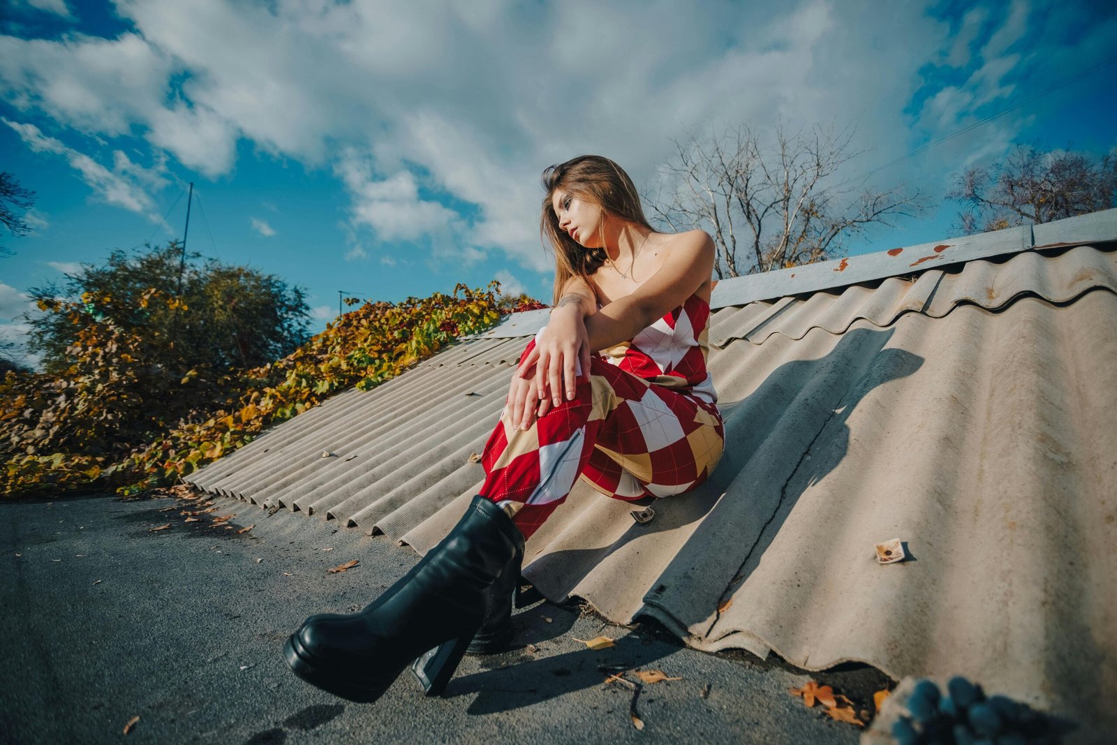 Stylish woman in harlequin outfit sits on rooftop under blue sky, embracing autumn vibes.